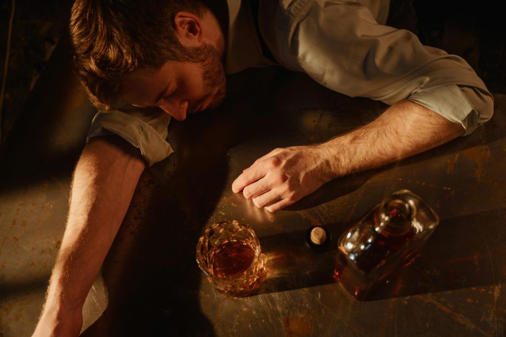 Un homme dort sur une table avec un verre et une bouteille de whisky à proximité, ce qui suggère l'épuisement.