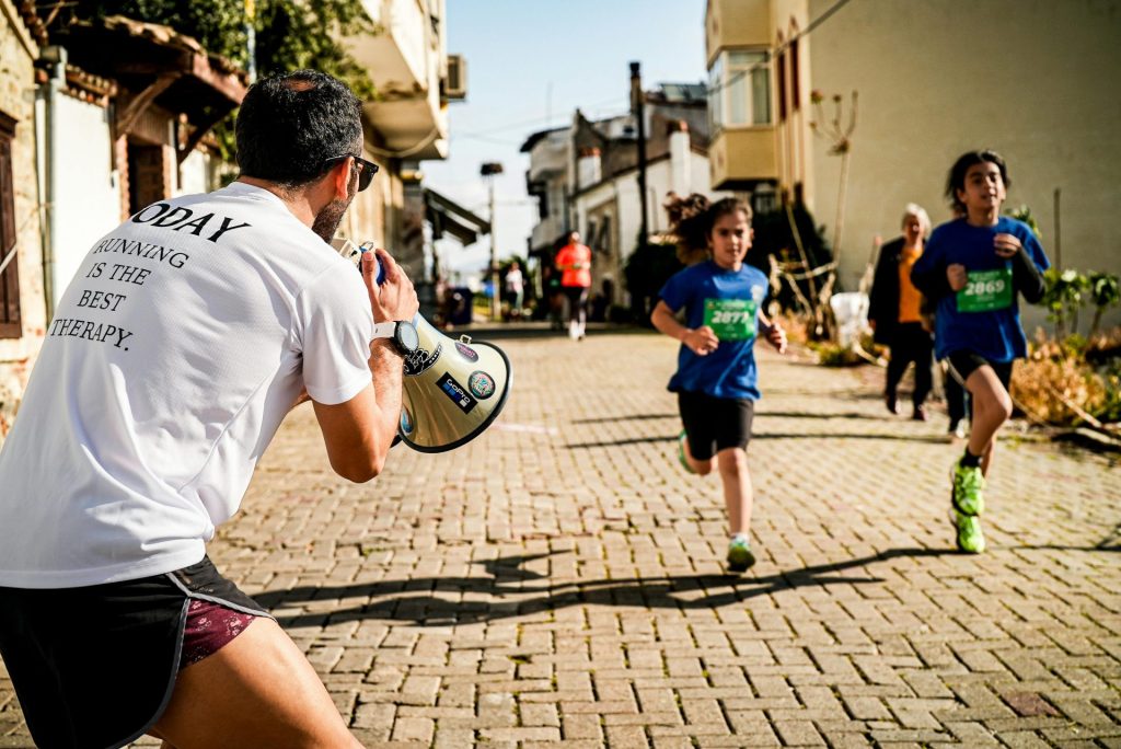 Des enfants participent à une course de rue tandis qu'un homme avec un mégaphone les encourage.