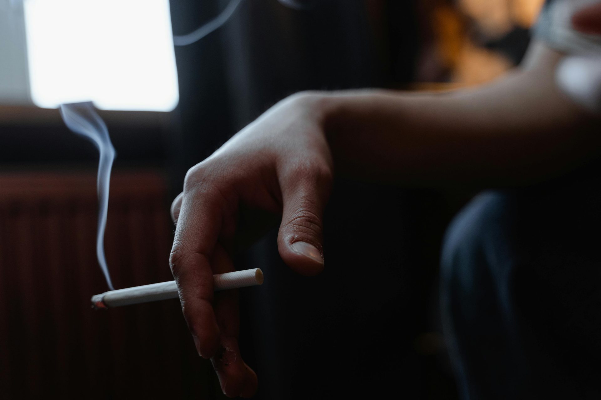 Gros plan d'une main tenant une cigarette allumée à l'intérieur avec de la fumée s'élevant dans une pièce faiblement éclairée.